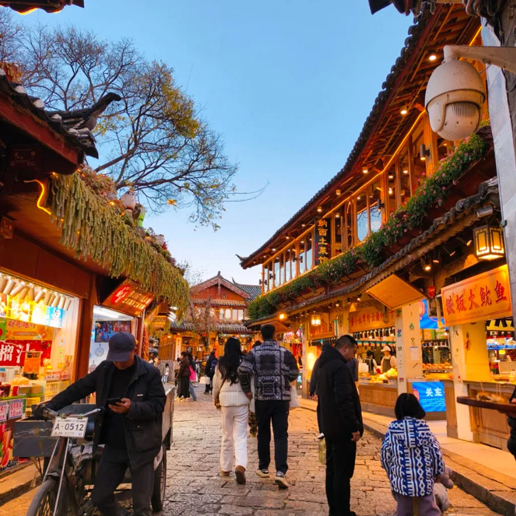 雲南著名歷史景區 麗江古城 的傍晚街景。石板路兩側是傳統木質建築與商店，屋簷掛著燈飾與招牌。遊客與當地居民在古街上漫步購物，街道燈光溫暖，營造出熱鬧又富有文化氣息的古鎮氛圍，展現麗江古城獨特的旅遊魅力