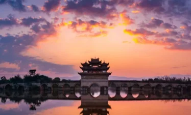 建水雙龍橋夕陽景觀，古老石拱橋橫跨水面，橋中央的傳統亭閣在晚霞映照下形成優美剪影，橋洞與天空雲彩倒映在平靜水面上，呈現雙龍橋典雅壯麗且充滿歷史韻味的古橋風光