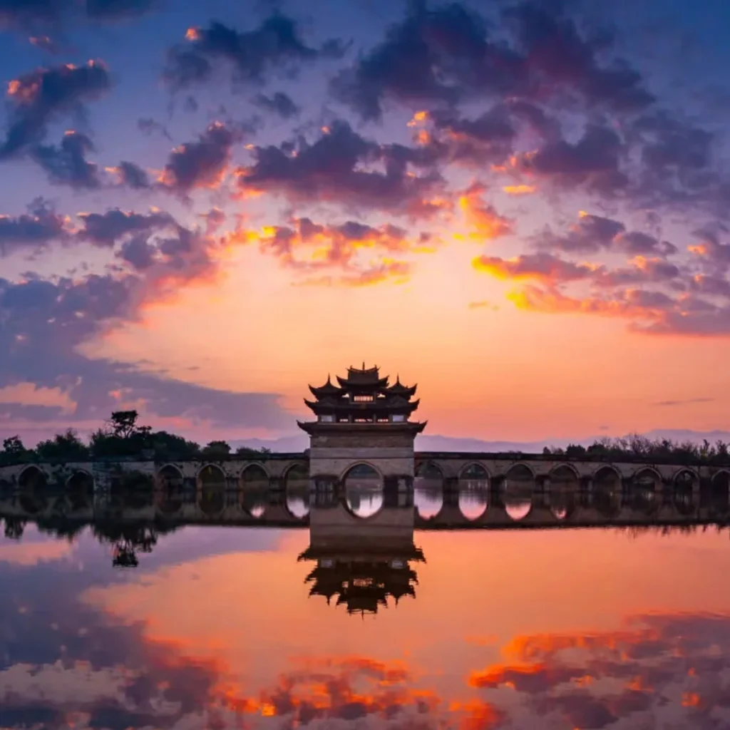 建水雙龍橋夕陽景觀，古老石拱橋橫跨水面，橋中央的傳統亭閣在晚霞映照下形成優美剪影，橋洞與天空雲彩倒映在平靜水面上，呈現雙龍橋典雅壯麗且充滿歷史韻味的古橋風光