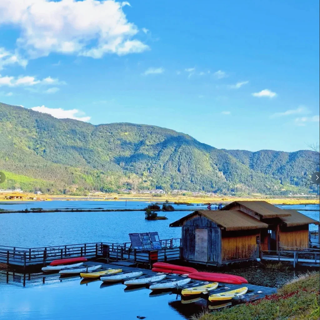 Tengchong Beihai Wetland 騰沖北海濕地自然景觀,湖面平靜映照藍天與群山,岸邊木屋與棧道延伸至水面,小船整齊停泊於碼頭旁。遠方青山環繞濕地湖泊,展現騰沖北海濕地獨特的高原湖泊生態與寧靜優美的自然風光