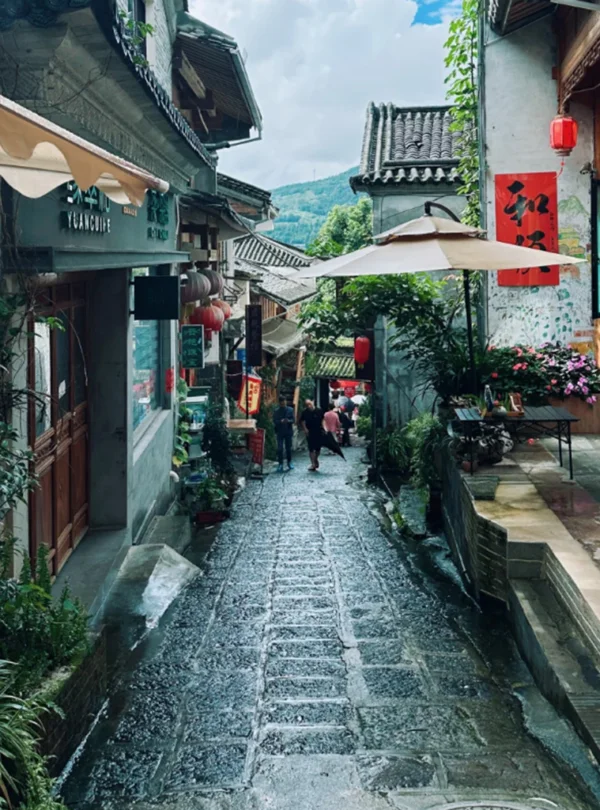 騰沖和順古鎮石板街巷景觀，兩側為傳統中式建築與特色店鋪，紅燈籠與庭院綠植點綴其間，遠處可見山景與遊客漫步其間，展現古鎮悠閒氛圍與濃厚歷史文化氣息。
