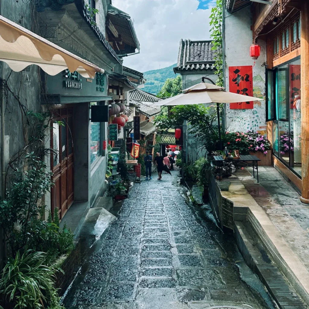 騰沖和順古鎮石板街巷景觀，兩側為傳統中式建築與特色店鋪，紅燈籠與庭院綠植點綴其間，遠處可見山景與遊客漫步其間，展現古鎮悠閒氛圍與濃厚歷史文化氣息。