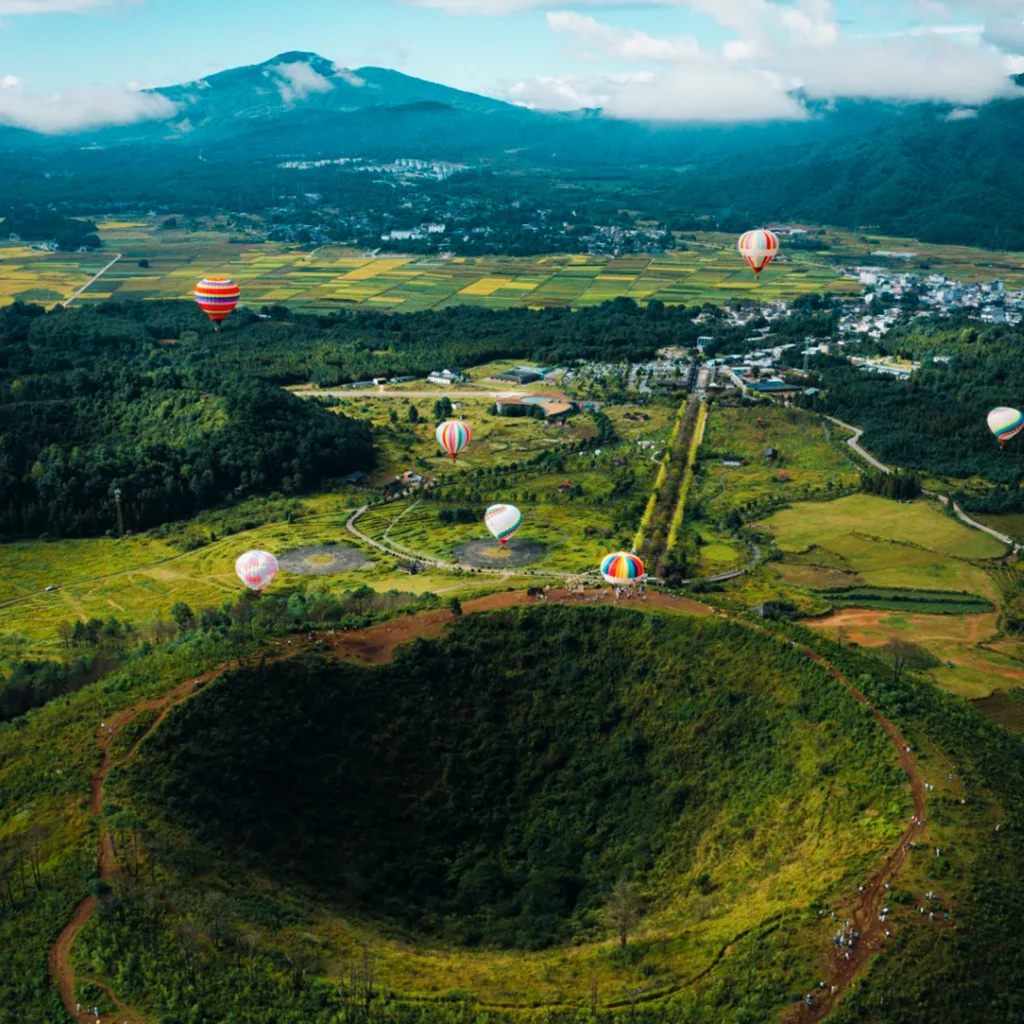 騰沖火山地熱國家地質公園壯觀火山口地形與熱氣球景觀，從高空俯瞰完整火山錐與綠色森林環繞，多彩熱氣球緩緩升空，遠處田園與山巒相連，展現騰沖火山地熱國家地質公園獨特火山地貌與自然生態景觀。