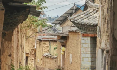 雲南團山民宅古村巷道景觀，狹窄石板路兩側是傳統土牆與瓦頂民居，老屋牆面帶有歲月痕跡，屋簷與綠植相互點綴，展現團山民宅保存完整的歷史建築與寧靜古村生活氛圍
