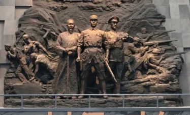 滇西抗戰紀念館大型歷史浮雕雕塑，呈現抗日戰爭時期軍民並肩作戰的場景，中央人物象徵指揮與守護家園的決心，上方飛機與戰士形象重現滇西戰場的激烈戰鬥與歷史記憶。
