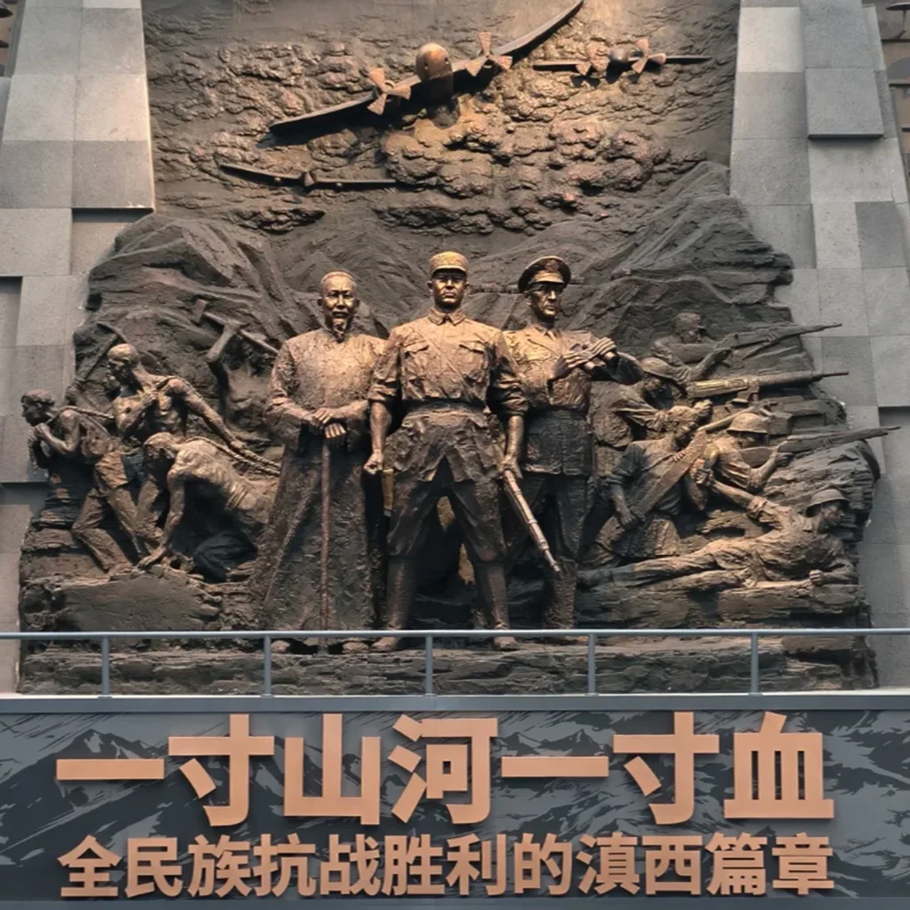 滇西抗戰紀念館大型歷史浮雕雕塑，呈現抗日戰爭時期軍民並肩作戰的場景，中央人物象徵指揮與守護家園的決心，上方飛機與戰士形象重現滇西戰場的激烈戰鬥與歷史記憶。