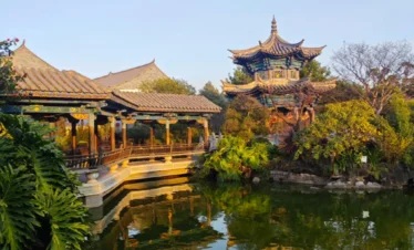 雲南著名古建築園林 朱家花園 的亭台水景。畫面中傳統中式長廊與雕梁畫棟的樓閣環繞池塘而建，金色夕陽照亮屋頂與木柱，綠樹與園林植物環繞，池水倒映古建築，展現典型中國古典園林景觀。