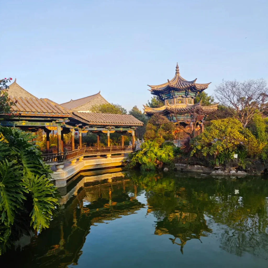 雲南著名古建築園林 朱家花園 的亭台水景。畫面中傳統中式長廊與雕梁畫棟的樓閣環繞池塘而建，金色夕陽照亮屋頂與木柱，綠樹與園林植物環繞，池水倒映古建築，展現典型中國古典園林景觀。