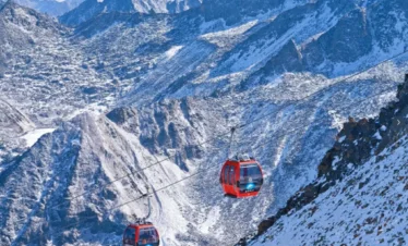 雲南麗江玉龍雪山冰川公園大索道的高山纜車景觀。紅色纜車在高空運行，穿越壯麗的雪山與冰川山脈，遠處連綿山峰覆蓋著白雪。崎嶇山脊與冰雪地貌展現高原壯觀景色，整個畫面呈現出震撼的自然風光與著名的高山旅遊體驗。