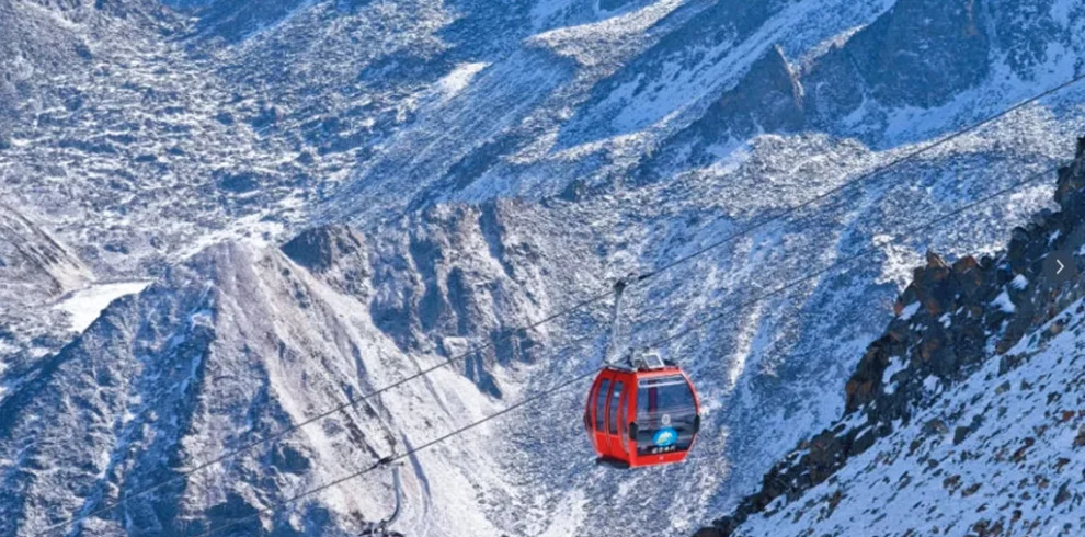 雲南麗江玉龍雪山冰川公園大索道的高山纜車景觀。紅色纜車在高空運行，穿越壯麗的雪山與冰川山脈，遠處連綿山峰覆蓋著白雪。崎嶇山脊與冰雪地貌展現高原壯觀景色，整個畫面呈現出震撼的自然風光與著名的高山旅遊體驗。