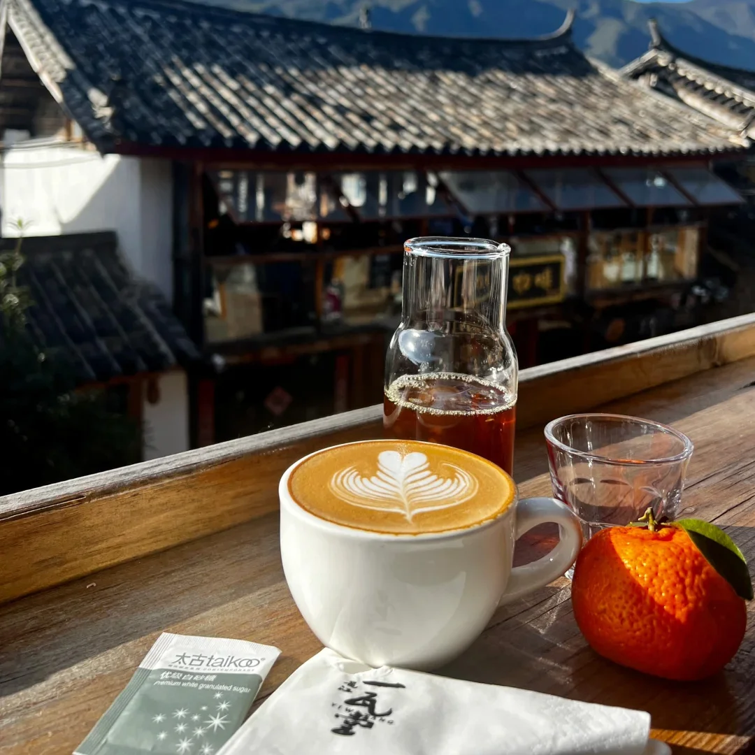 麗江古城小巷咖啡店的愜意咖啡時光。拉花拿鐵、冰茶、新鮮橘子，陽光下襯著傳統納西族建築。