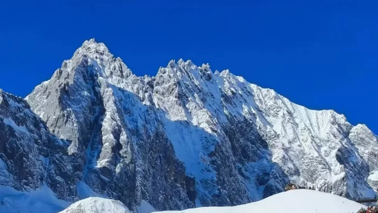 雲南麗江玉龍雪山，雄偉鋸齒狀積雪山峰聳立於澄澈深藍天空下，山腳有小型觀景平台。