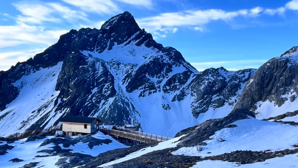 雲南麗江玉龍雪山，雄偉積雪山峰拔地而起，山腳有木棧道與山中小屋，背景是澄澈藍天。