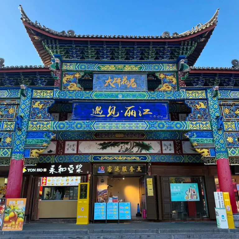 雲南麗江古城大研花巷，雕梁畫棟的傳統中式牌樓，「遇見・麗江」字樣，下方是在地店家，背景是澄澈藍天。