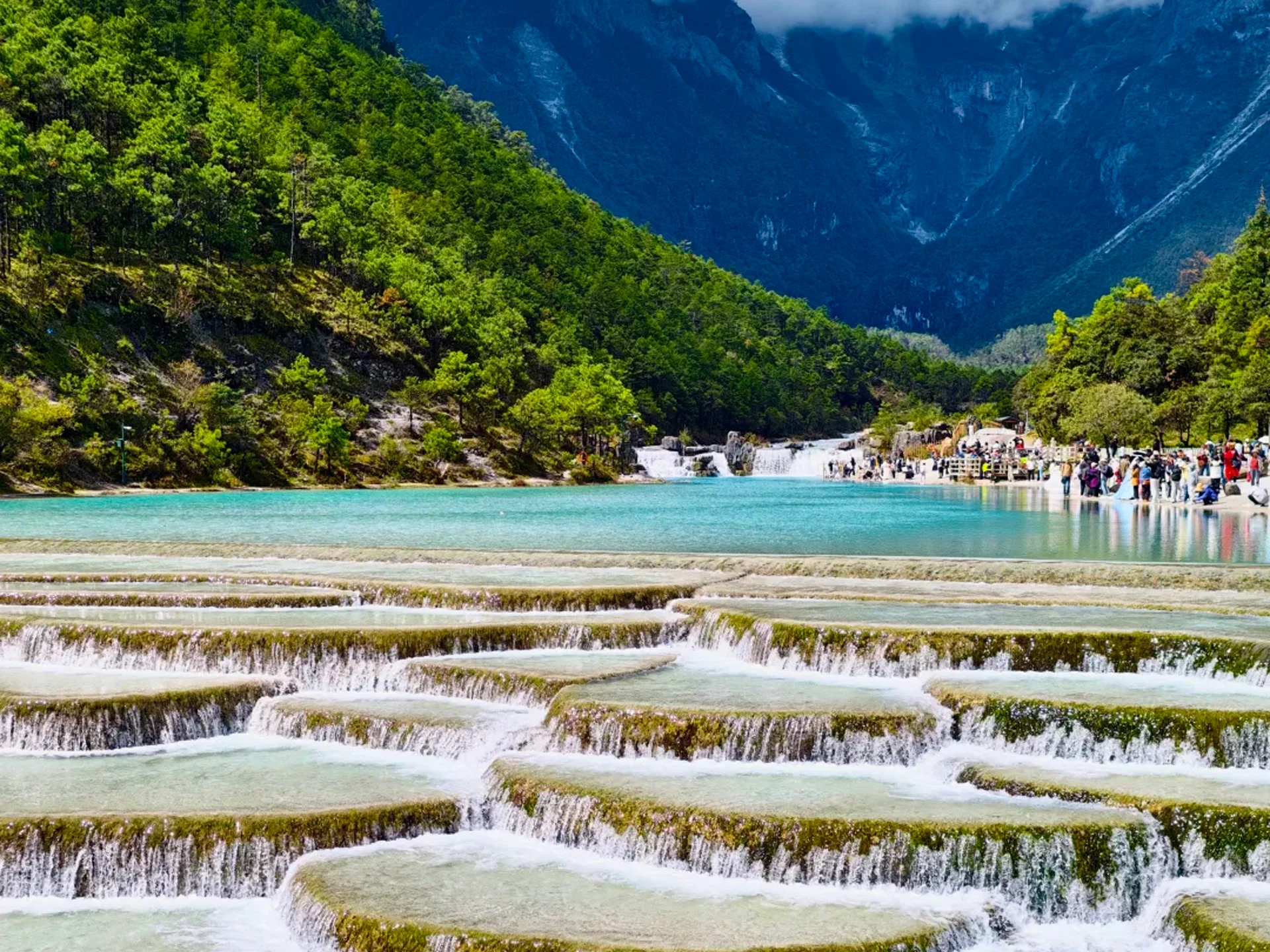 雲南麗江白水台，層層疊疊的鈣華階梯瀑布流淌著蒂芬妮藍湖水，兩側是蒼鬱山林，遠處環繞巍峨雪山，宛如人間仙境。