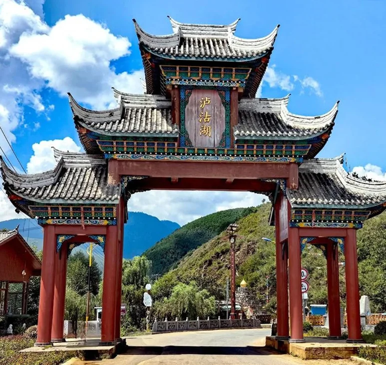 Lugu Lake 1 瀘沽湖景區入口的傳統中式牌樓矗立在道路上方,紅色立柱與多層飛簷屋頂展現典雅建築風格。牌樓中央寫著「瀘沽湖」三字,背後是綠色山巒與藍天白雲,呈現通往瀘沽湖自然風景區的地標入口與濃厚地方特色。