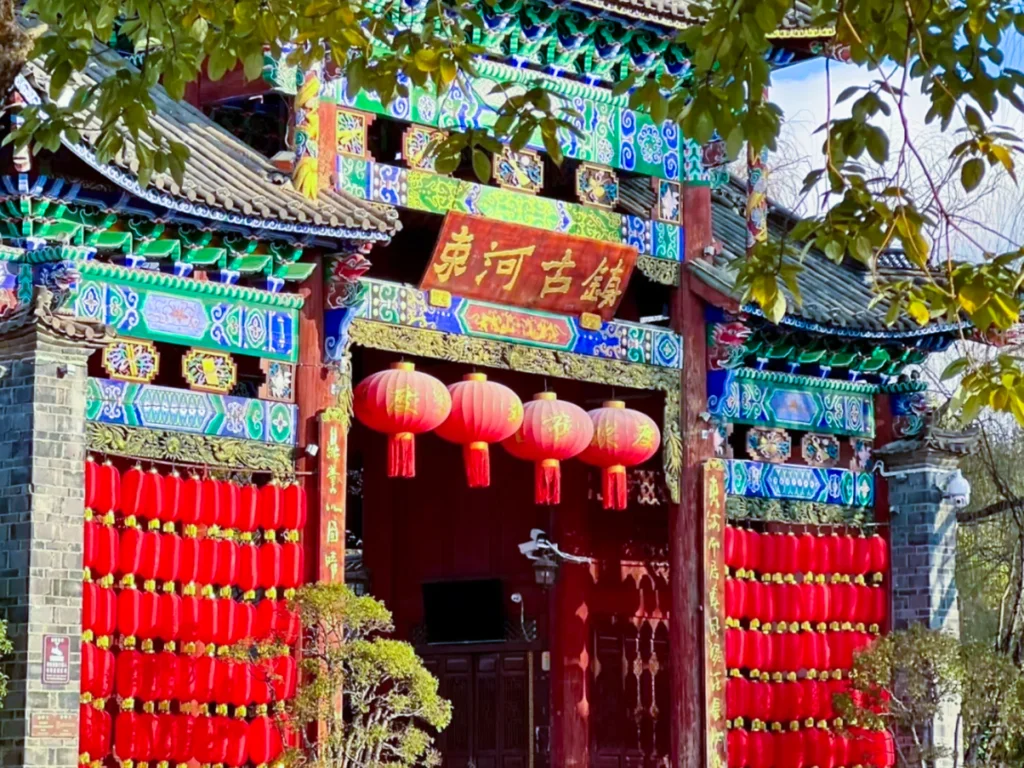 麗江束河古鎮入口，雕梁畫棟的傳統中式牌樓，掛滿紅燈籠與「束河古鎮」牌匾，背景是澄澈藍天。
