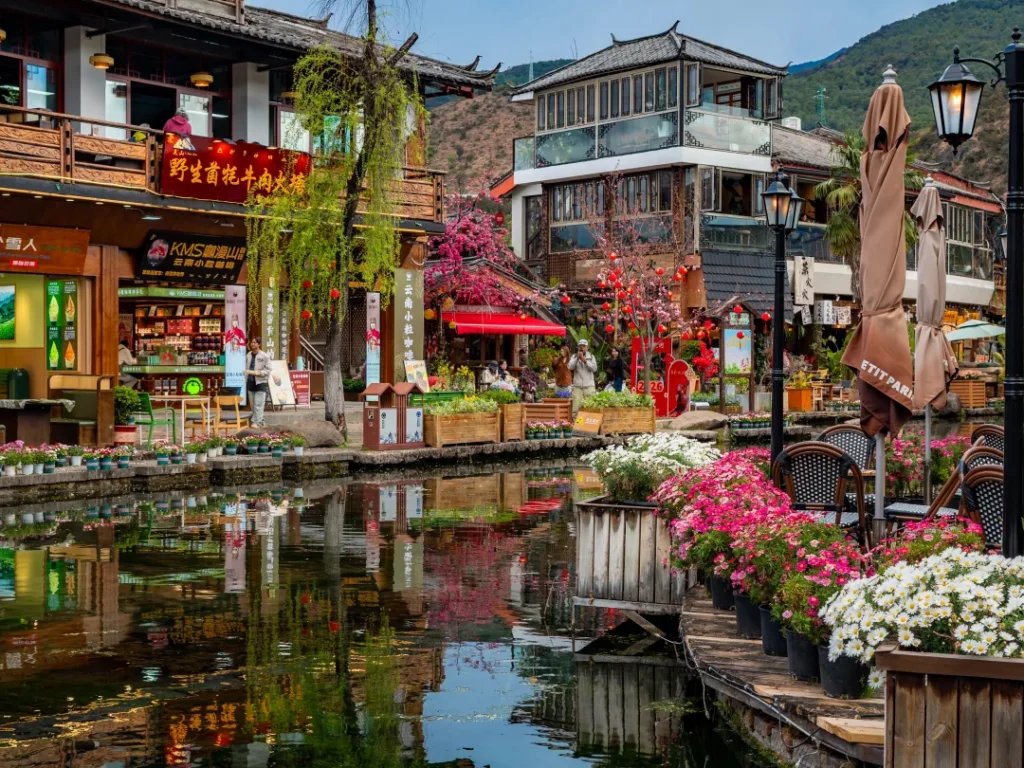 麗江束河古鎮的唯美水景，碧綠潭水倒映著翠綠垂柳、鮮豔花卉與古色古香的納西建築，呈現如畫般的夢幻景致。
