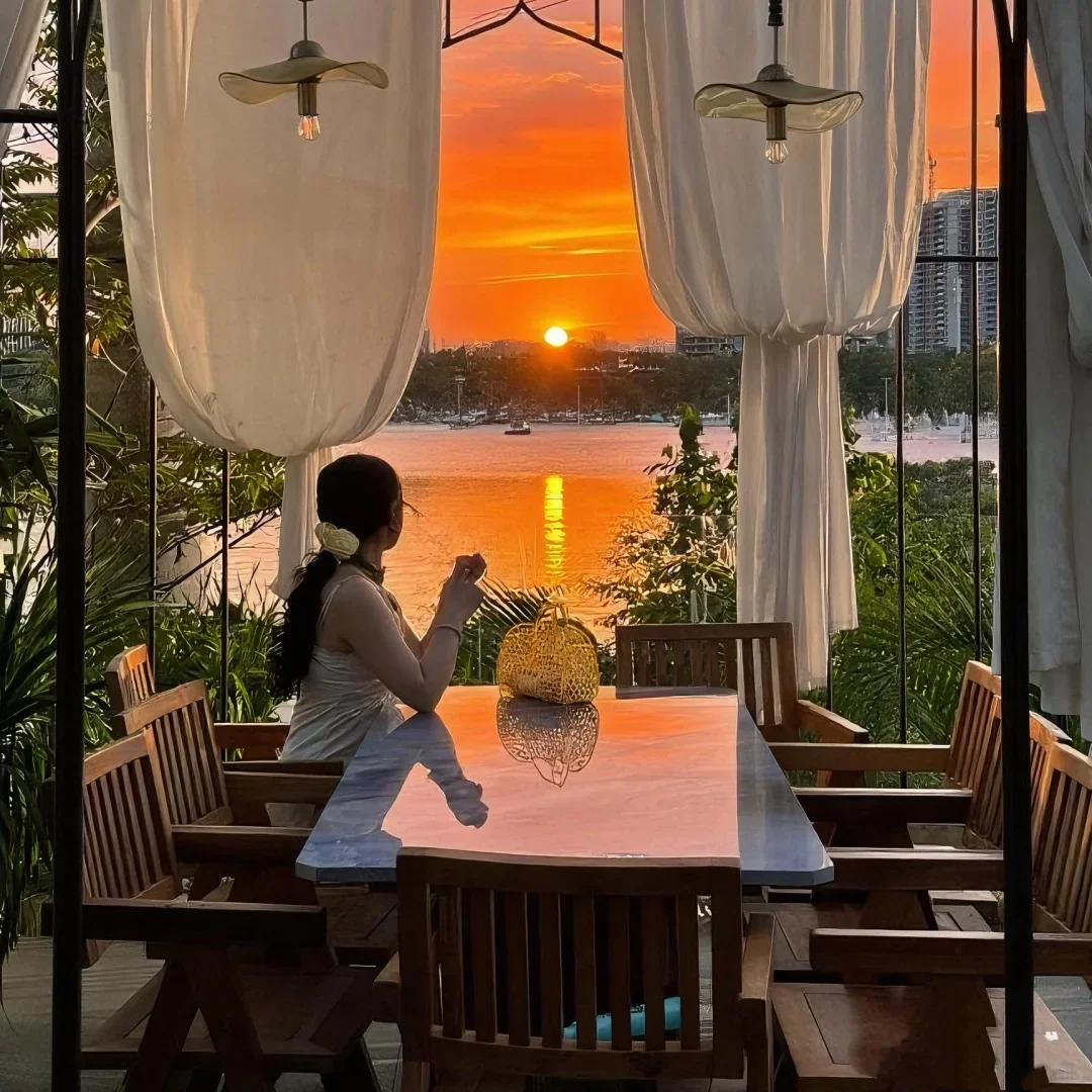 西雙版納湄公河畔咖啡店的露天座位，一名女子獨自坐在長桌前凝望壯麗的夕陽，金紅色的陽光倒映在江面與大理石桌上。