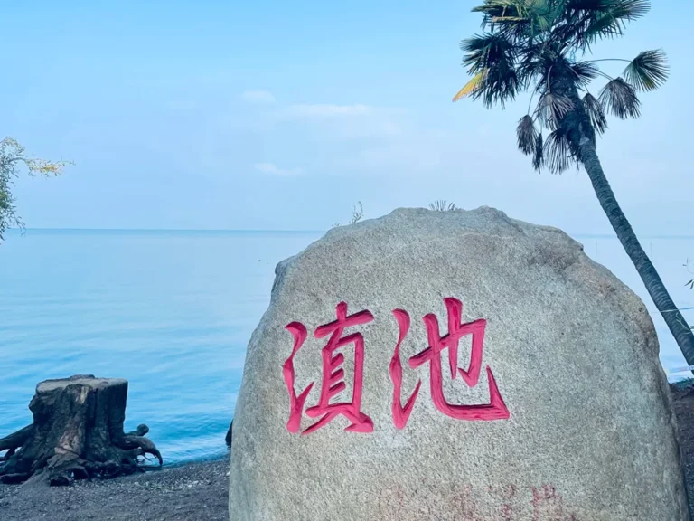 昆明滇池的經典地標石碑，刻有紅色「滇池」大字的灰色石頭，背景是湛藍的湖水與藍天，旁邊還有棕櫚樹點綴。