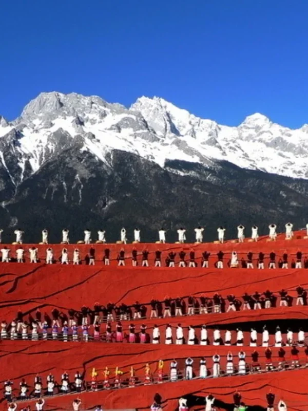 印象麗江大型實景表演在玉龍雪山下上演，紅色階梯舞台上眾多演員身著民族服飾整齊排列演出，背後是壯麗的雪山與藍天景觀，展現麗江獨特的民族文化與印象麗江震撼的戶外表演場面