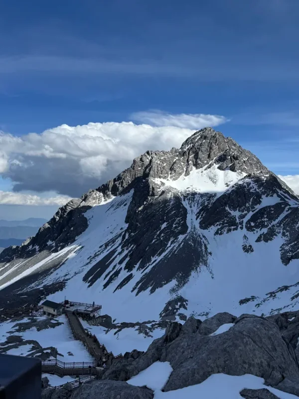 雲南麗江玉龍雪山，雄偉雪山聳立於澄澈藍天之下，木棧道沿著岩坡蜿蜒向上，盡顯高原壯麗風光。