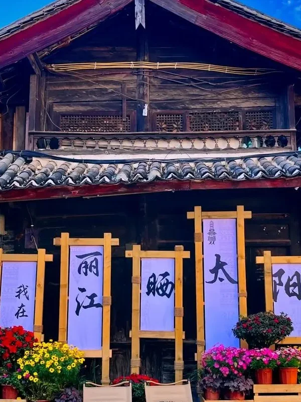 麗江古城傳統木造建築外觀，屋頂覆蓋灰色瓦片，展現納西族古城建築特色。門前擺放花卉盆栽與木製展示架，上方書寫「我在麗江晒太陽」字樣，呈現悠閒的古城生活氛圍，是遊客拍照與體驗麗江古城文化的熱門場景。