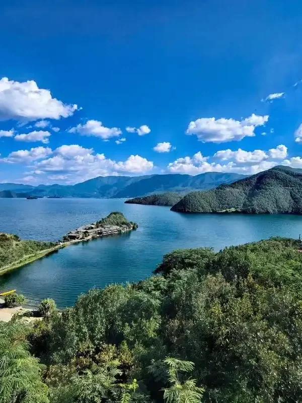 瀘沽湖壯麗湖景在晴朗藍天下展開，湖水深藍清澈，群山環繞湖泊。湖中央的小島與連接岸邊的堤道清晰可見，周圍綠意盎然的森林與遠方山巒層層延伸，白雲點綴天空，展現瀘沽湖高原湖泊寧靜而壯觀的自然風光。