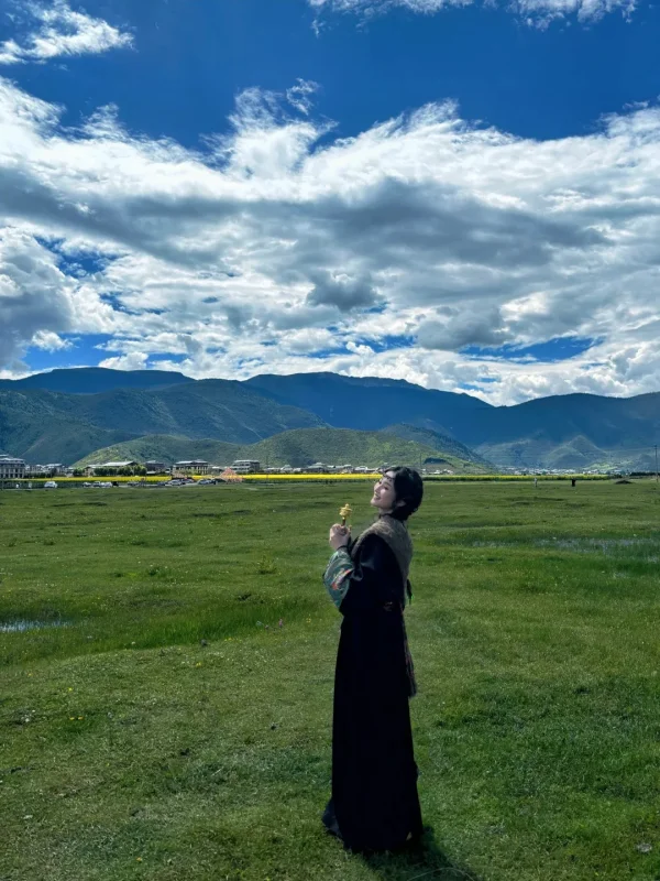 Shangri La Napahai 3 在納帕海草原上,穿著藏服、手持轉經輪,背後是遼闊的草甸、遠山與藍天白雲,是香格里拉最具代表性的旅行寫真畫面。