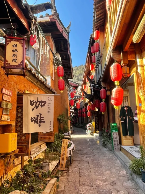 束河古鎮的陽光古巷，紅燈籠高掛、木質建築溫暖，藍天下的青石板路，是麗江最具慢生活氛圍的巷弄畫面。