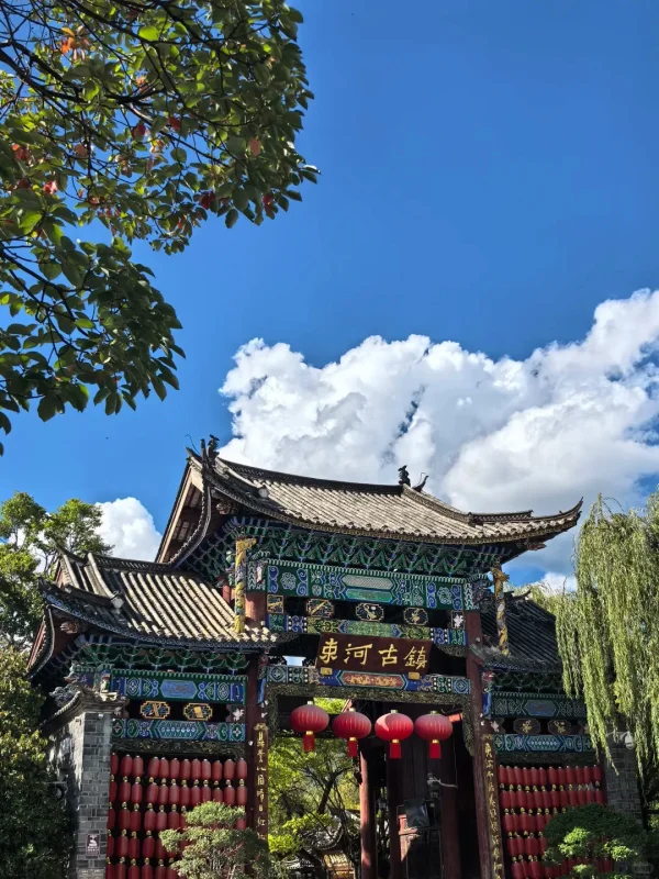 麗江束河古鎮的牌樓，彩繪木雕與紅燈籠相映，藍天白雲下，是古鎮最具儀式感的入口畫面。